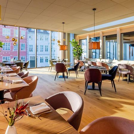 Nh Collection Copenhagen Otel Dış mekan fotoğraf The photo depicts a modern restaurant interior with ample natural light. The space features large windows that reveal buildings outside, enhancing the ambiance. Inside, there are several dining tables arranged with elegant table settings, including c